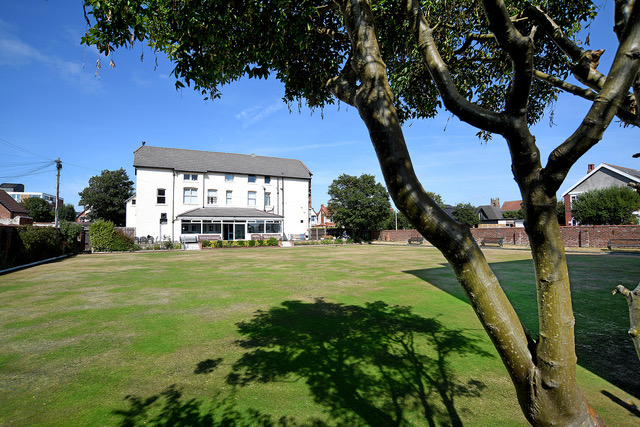 The bowling green from beneath a mature tree, the white clubhouse rising behind the far edge on a bright summer day.