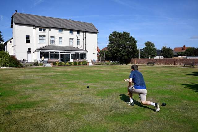 A bowler in a club polo shirt kneeling on the green as he delivers a wood, the clubhouse conservatory behind him.