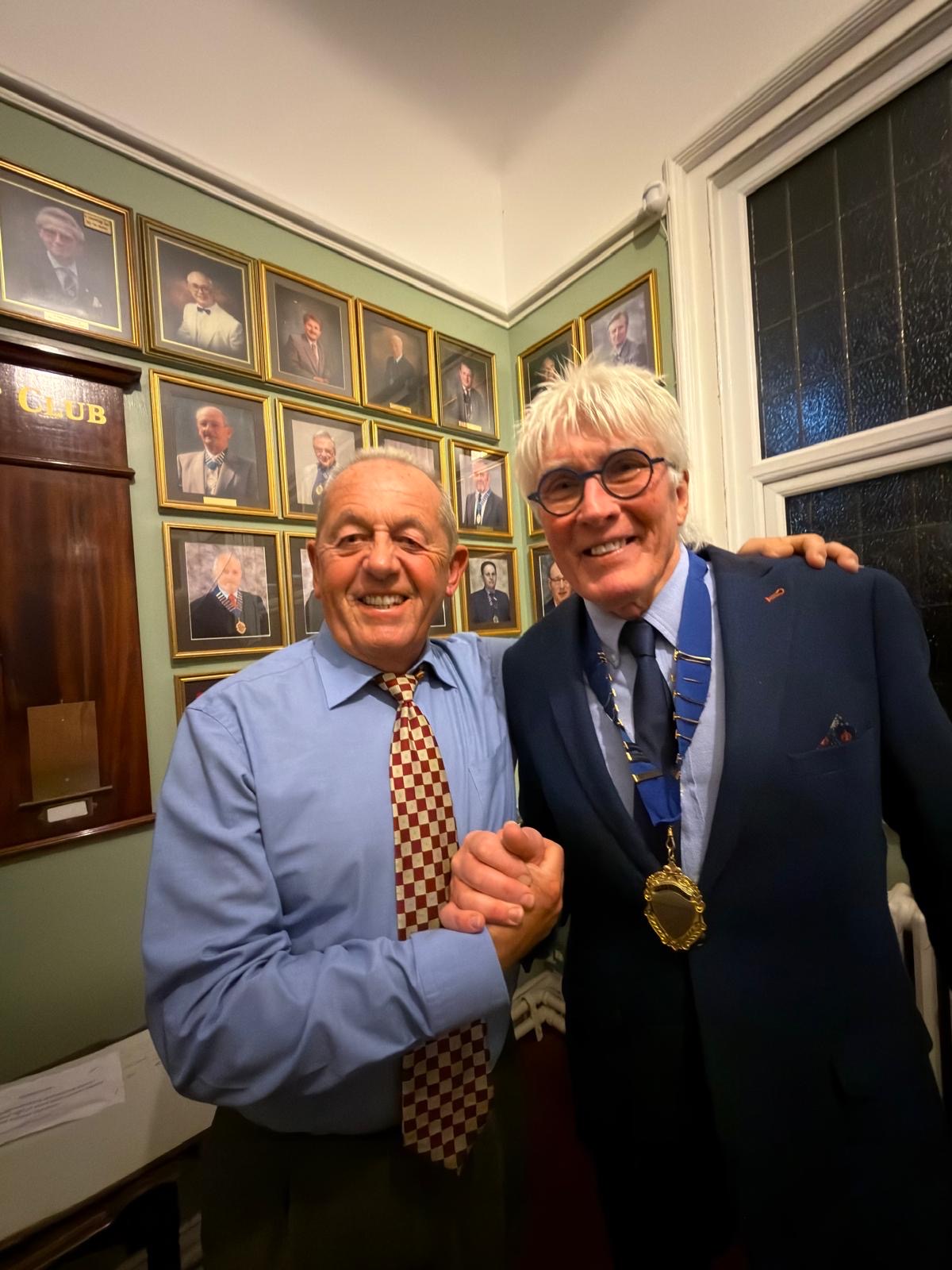 Two members shaking hands in front of the club's wall of framed past-president portraits, one wearing the chain of office.