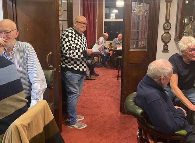 Members of The District Club in the lounge of an evening, one seated in a checked jumper reading a newspaper.